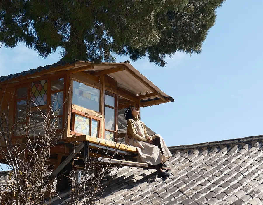 Wenjun sitting beside a wooden house on a tiled roof
